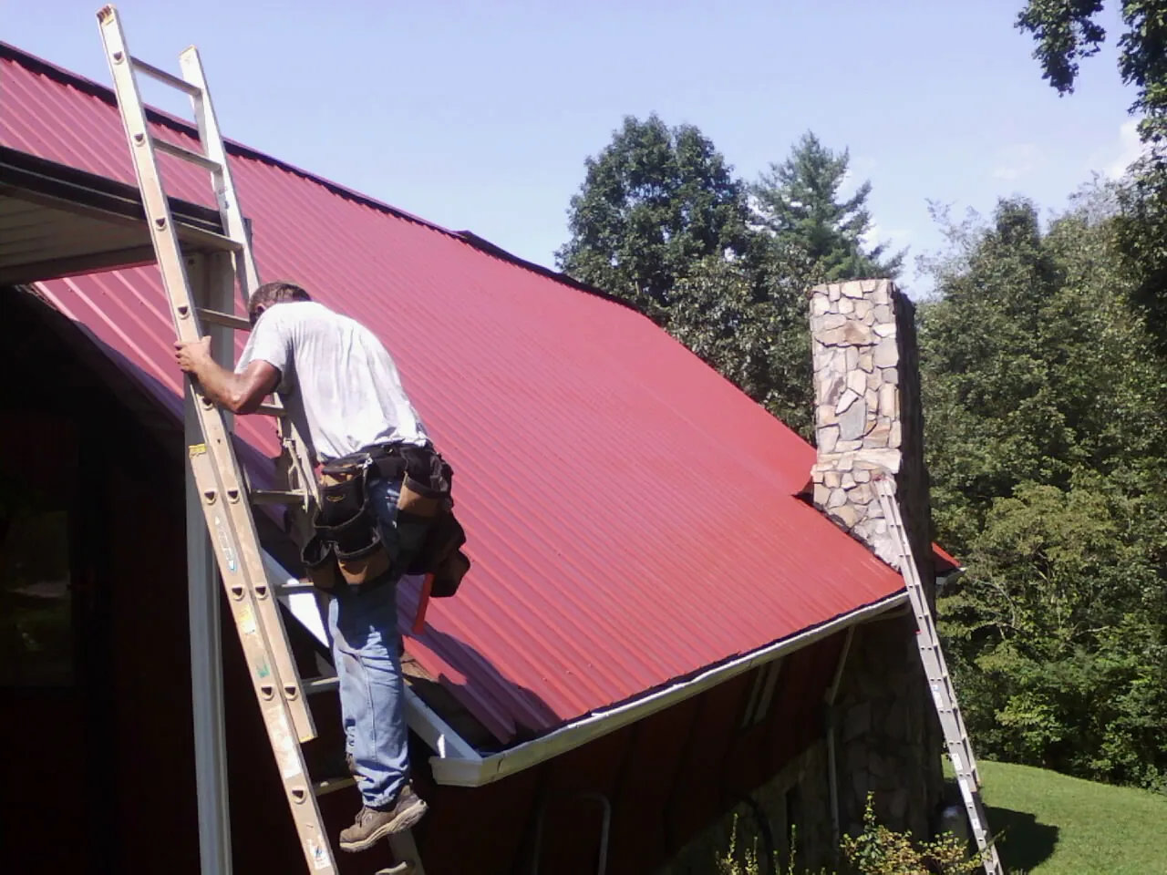 Roofer on a ladder beside a red metal roof and stone chimney — Soffit & Fascia Repair by Roofing Brothers in Methuen